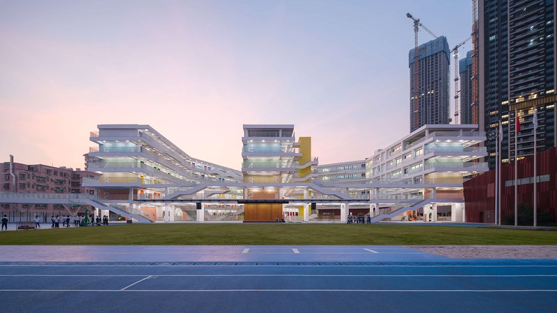Layered white facade with yellow accent tower viewed across a blue running track at dusk