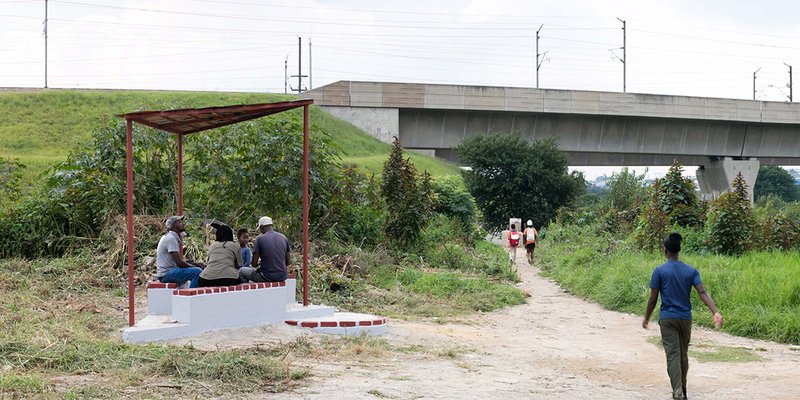 Shelter with undulating canopy on a dirt path with visitors walking toward a concrete highway overpass