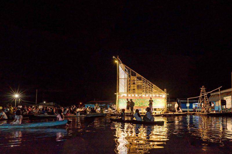 Illuminated bamboo lattice screen and projection wall reflected in dark water with boats nearby at night