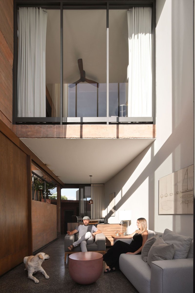 Double-height living room with cantilevered mezzanine bedroom and residents seated below a ceiling fan