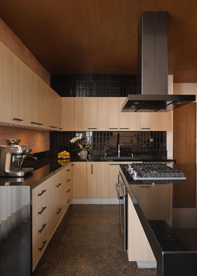 Galley kitchen with pale timber cabinetry, black tile backsplash and terrazzo flooring under warm ceiling