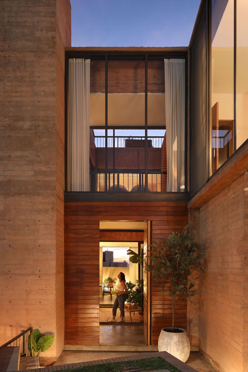 Courtyard entry with timber-clad facade and illuminated doorway framed by rammed earth walls at twilight