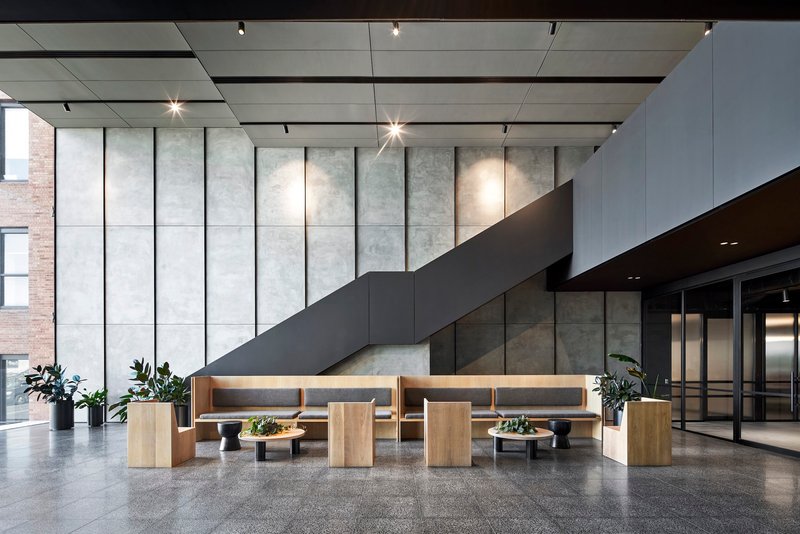Double-height lobby with gridded concrete wall panels, overhead track lighting, and timber seating banquettes below cantilevered staircase