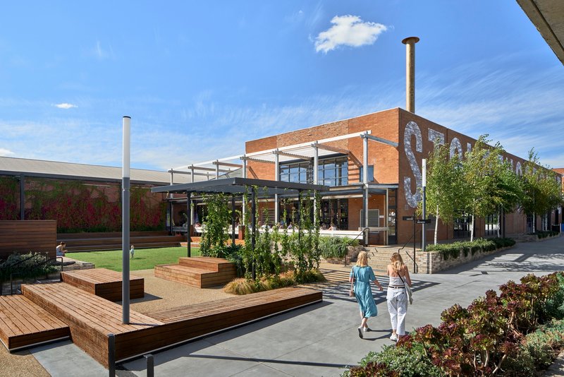 Courtyard with timber-edged planters and steel-framed canopy structures beneath a blue sky with scattered clouds