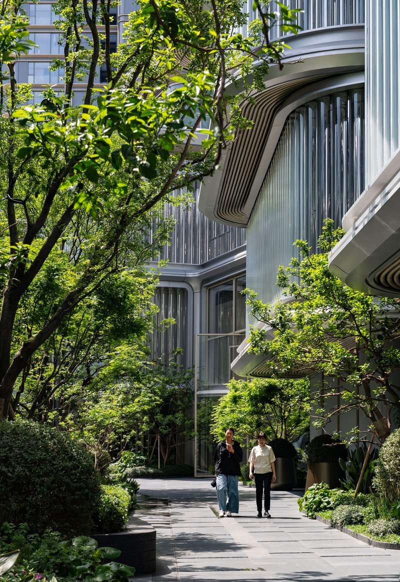 Curved facade with vertical metal cladding framed by mature trees along a landscaped pedestrian path