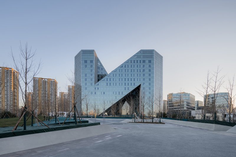 Zigzag glass curtain wall facade viewed from a paved plaza with bare winter trees