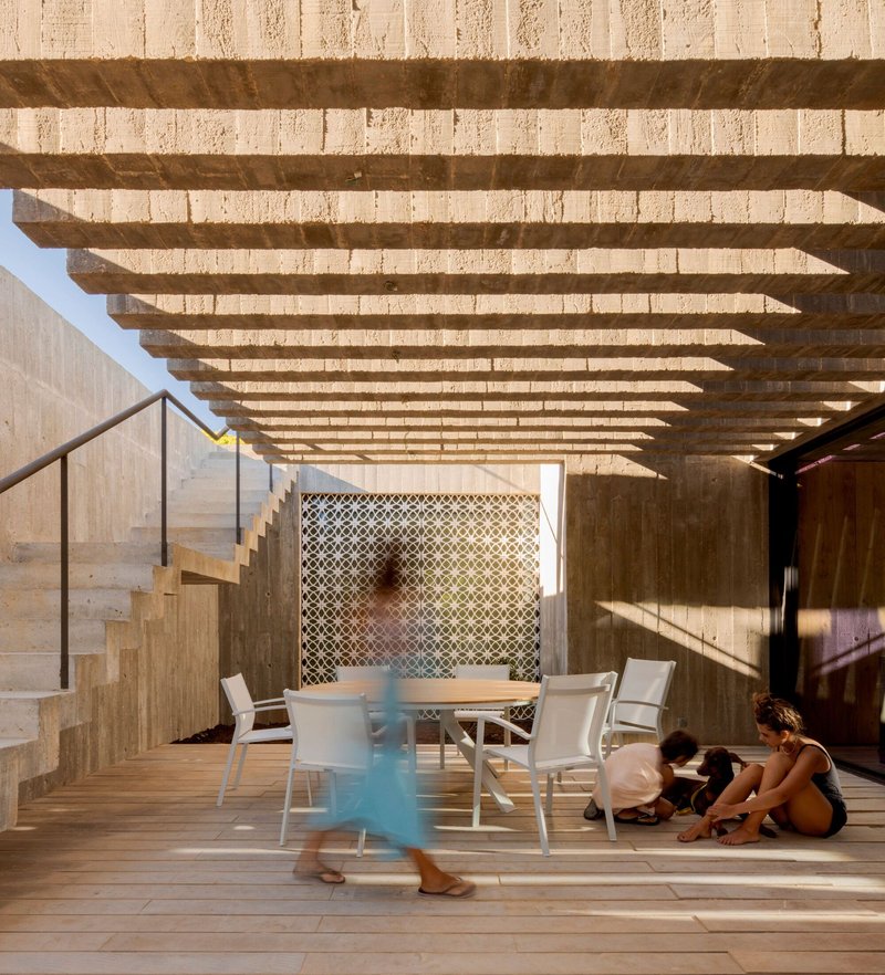 Covered terrace with timber pergola beams casting shadows over wooden decking and playing children