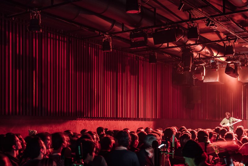 Performance space with red curtain backdrop and exposed technical rigging above a gathered audience