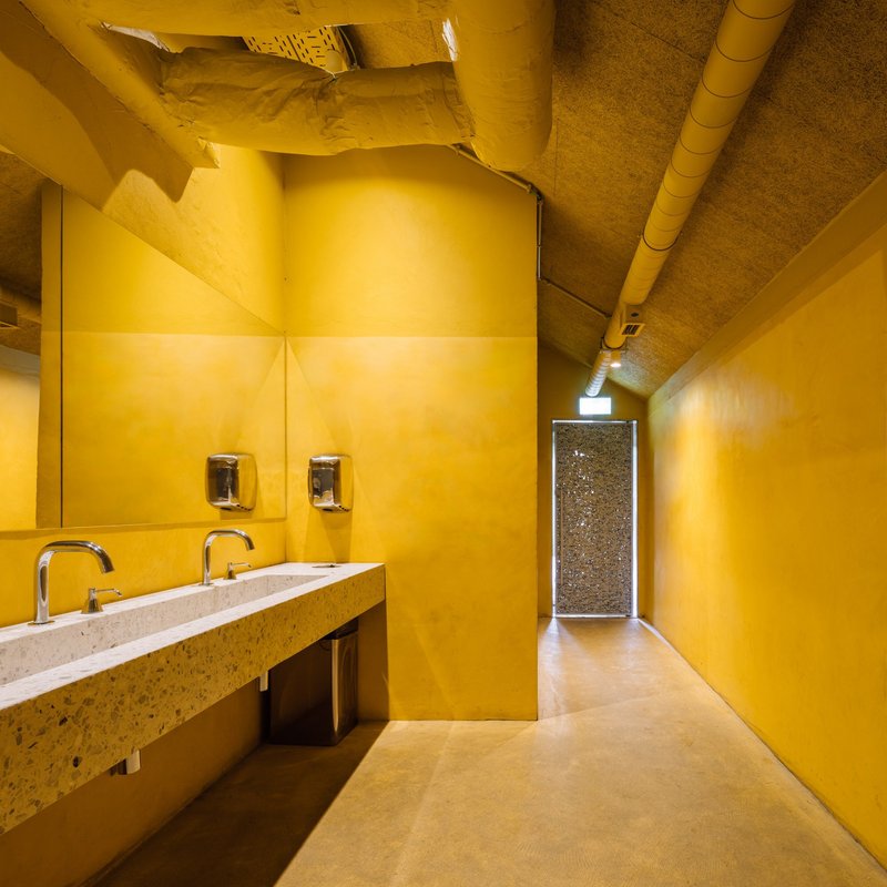 Restroom corridor with yellow-painted walls, terrazzo sink counter, exposed ductwork, and mirrored doorway at the end