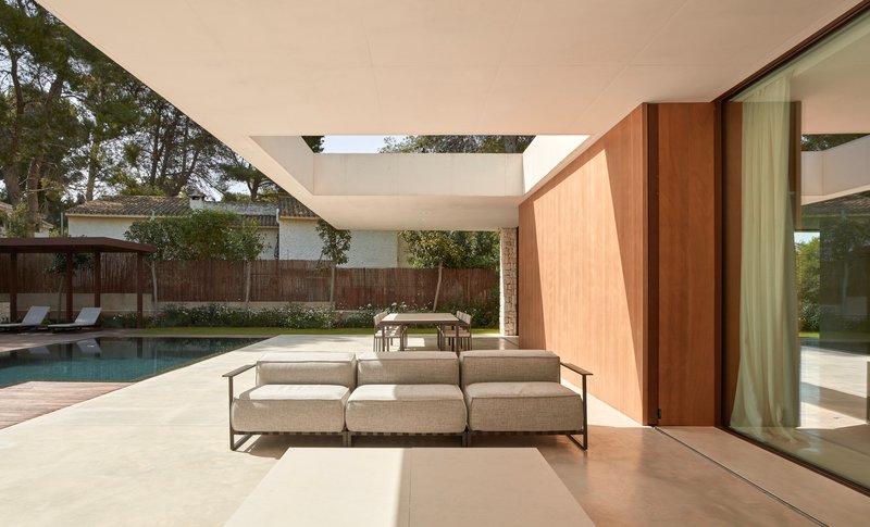Covered outdoor terrace with wood-paneled pivot door and low sofa overlooking a pool and timber fence