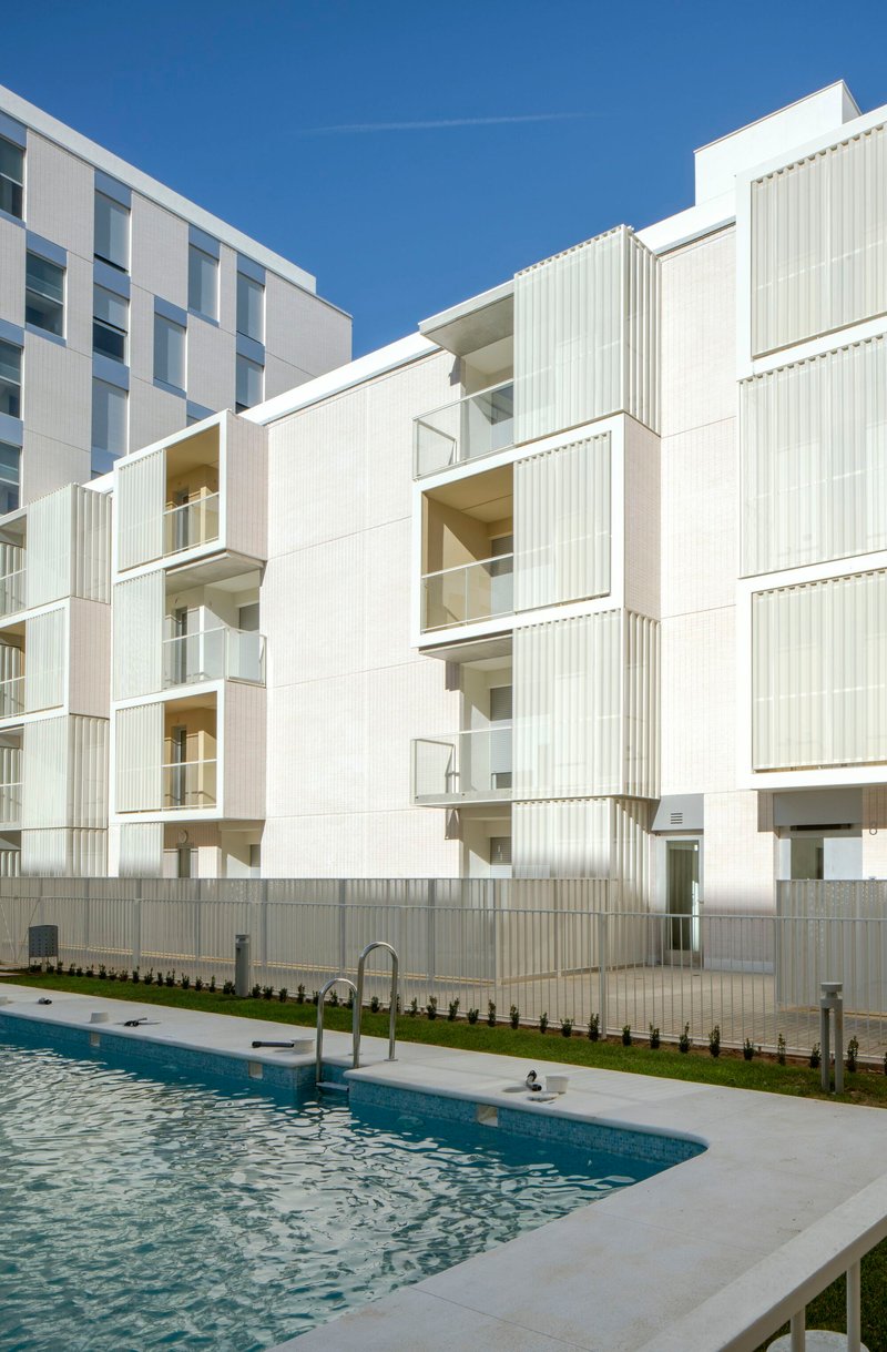 White facade with protruding balconies and vertical screening panels facing a swimming pool under blue sky