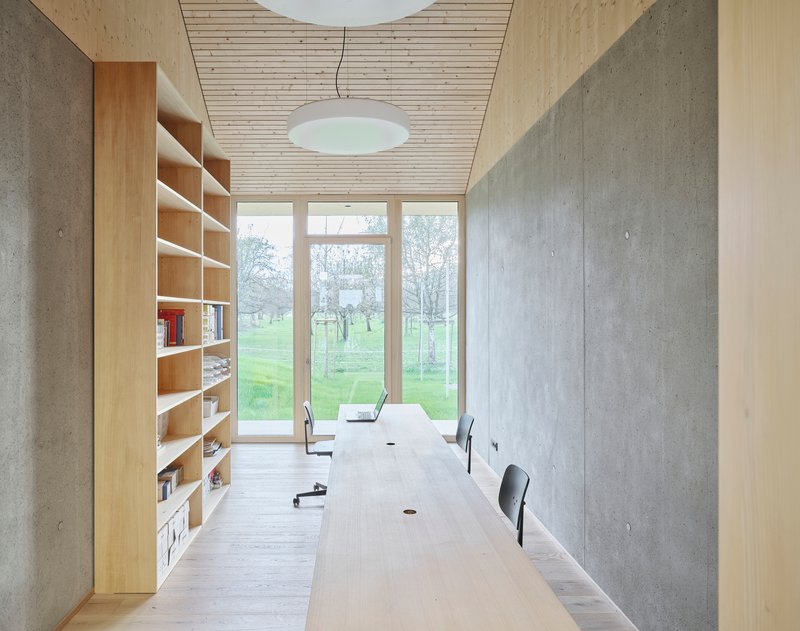 Long interior workroom with plywood shelving, concrete wall, timber table and disc pendant above