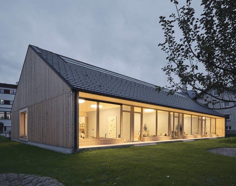 Corner view of the pitched-roof volume with illuminated glazed facade at dusk