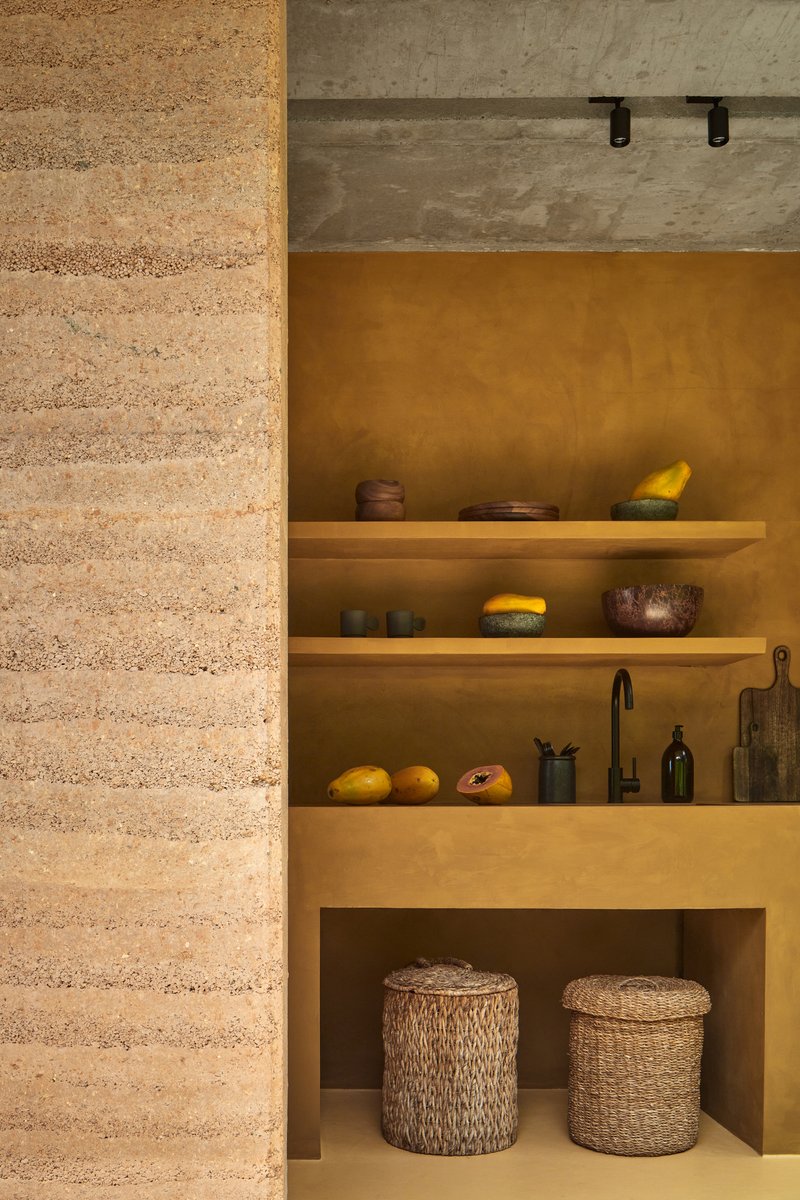 Kitchen alcove with floating timber shelves against ochre plaster walls beside a rammed earth partition