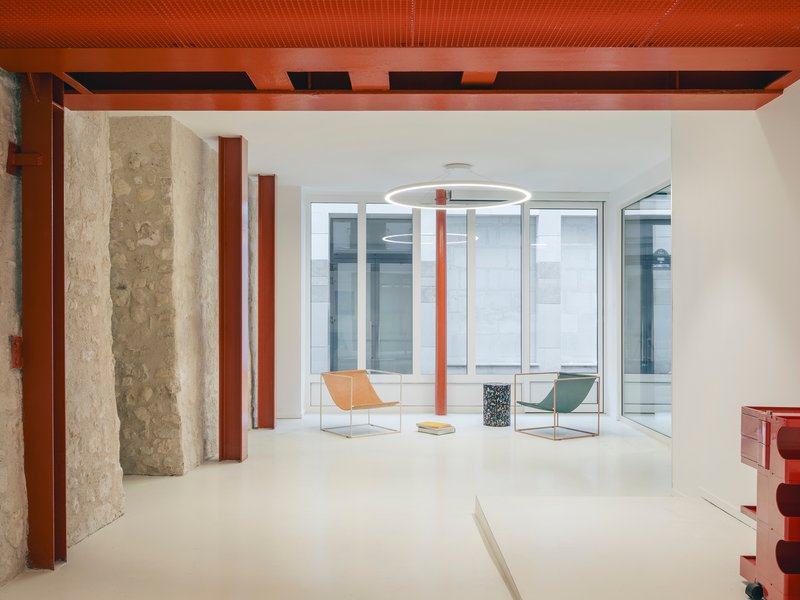 Interior space with exposed stone walls, red steel columns, perforated metal ceiling and glazed courtyard beyond