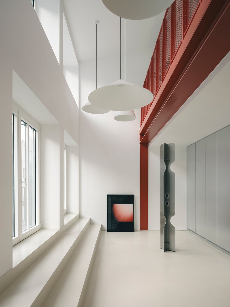 Double-height interior with red perforated metal mezzanine, pendant lights, and sunlight through tall windows