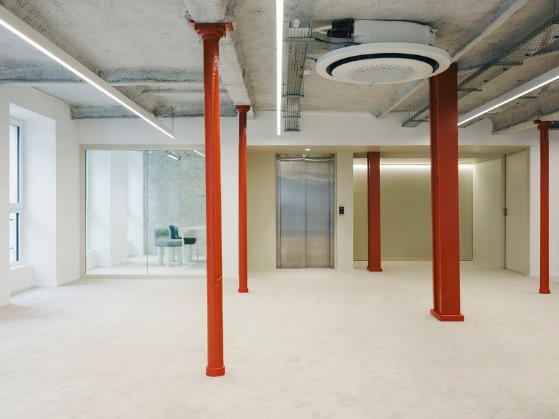 Open floor plan with exposed concrete ceiling, red columns, central elevator core and glass-walled meeting room
