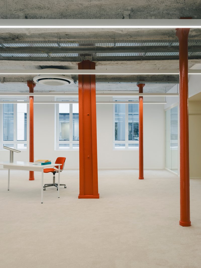 Orange steel columns framing workspace with desk and chair beneath exposed concrete ceiling and ducting