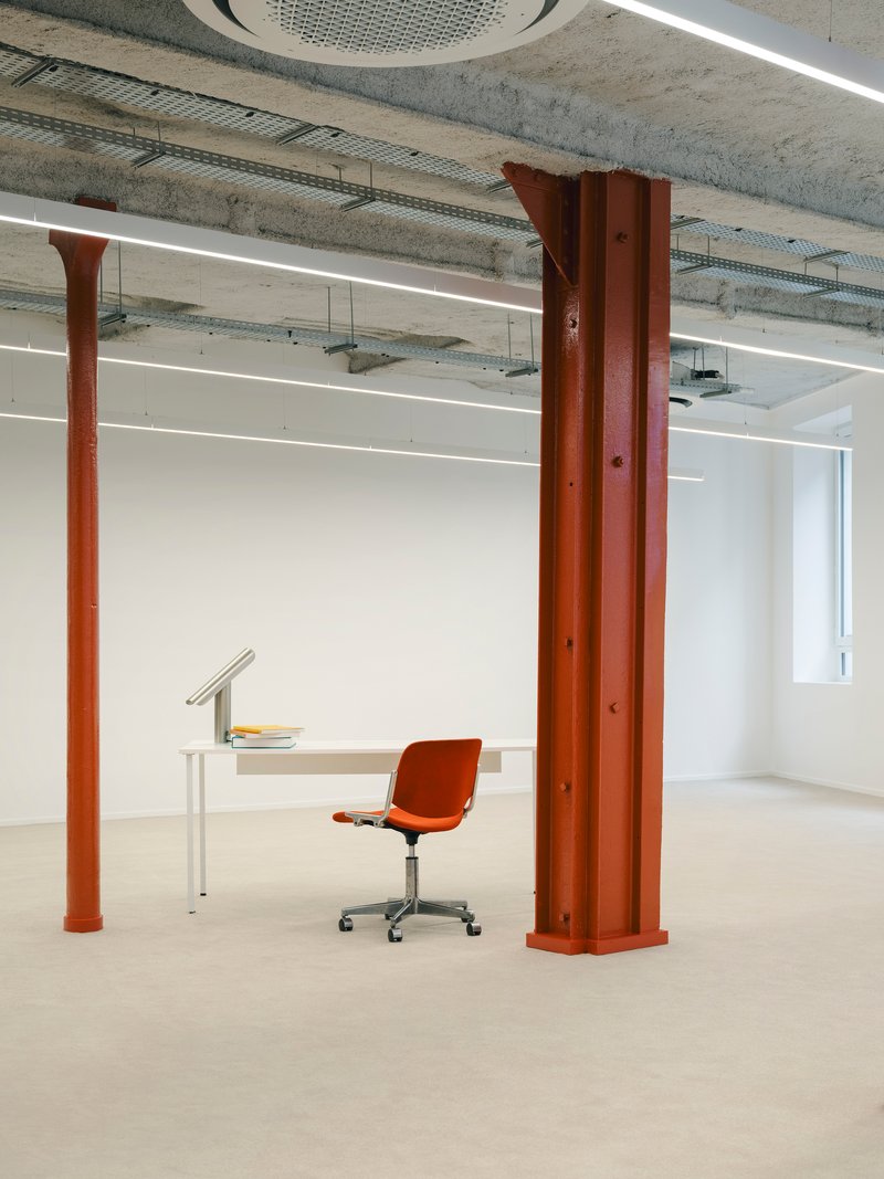 Orange steel column beside minimal white desk and chair beneath exposed concrete ceiling with linear lighting