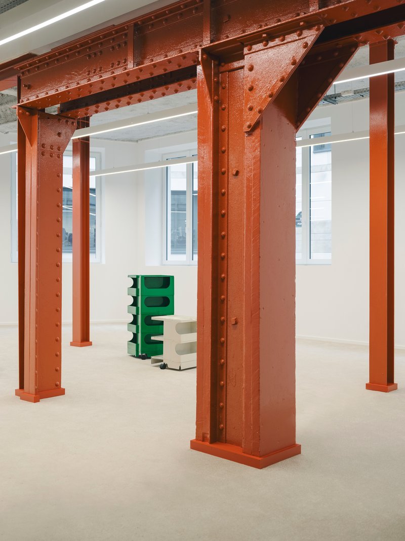 Exposed riveted steel beams and columns painted red oxide in an open floor space with white walls