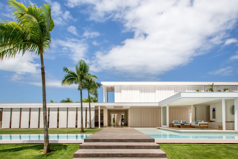 View across the lawn and pool deck toward the white volume with vertical louvers and palm trees