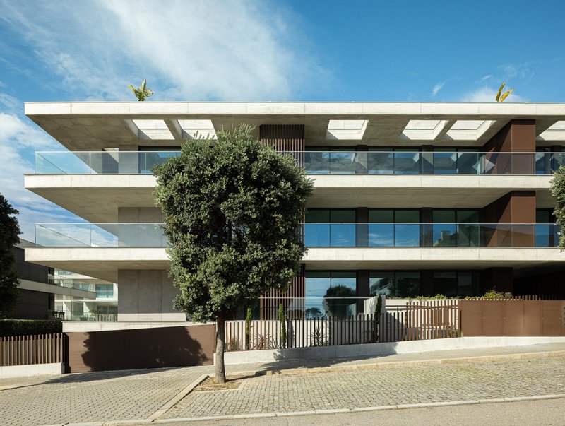 Street facade with cantilevered concrete balconies and glass railings framed by mature trees under blue sky