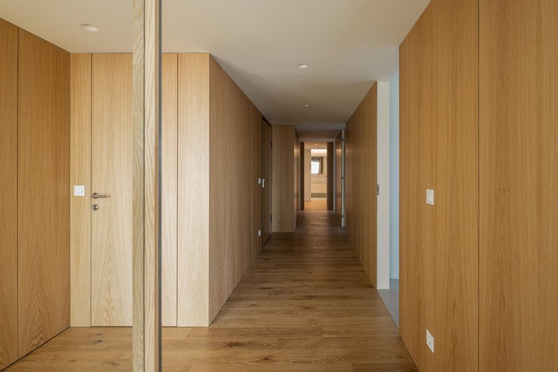 Long corridor lined with continuous timber wall paneling and flush doors extending toward a sunlit end