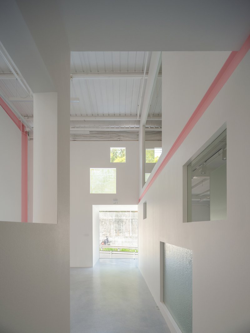 Corridor view through white partition walls with pink diagonal beams and stacked windows framing green foliage outside