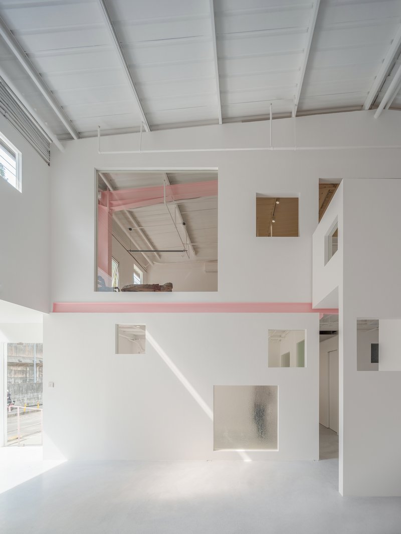 Double-height interior with white partition walls punctured by square openings and pink beam spanning upper mezzanine level