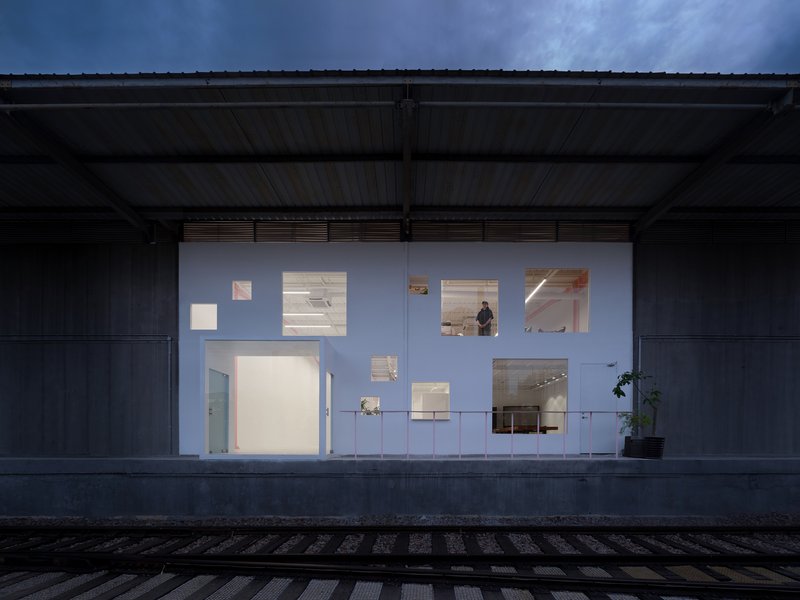 Illuminated white facade wall beneath a metal shed canopy at dusk with a figure visible inside