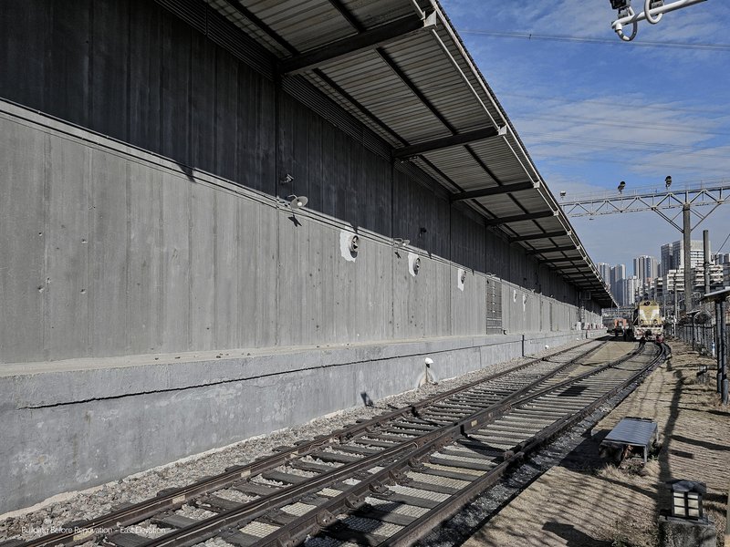 Long concrete wall with metal canopy running parallel to railway tracks under clear blue sky