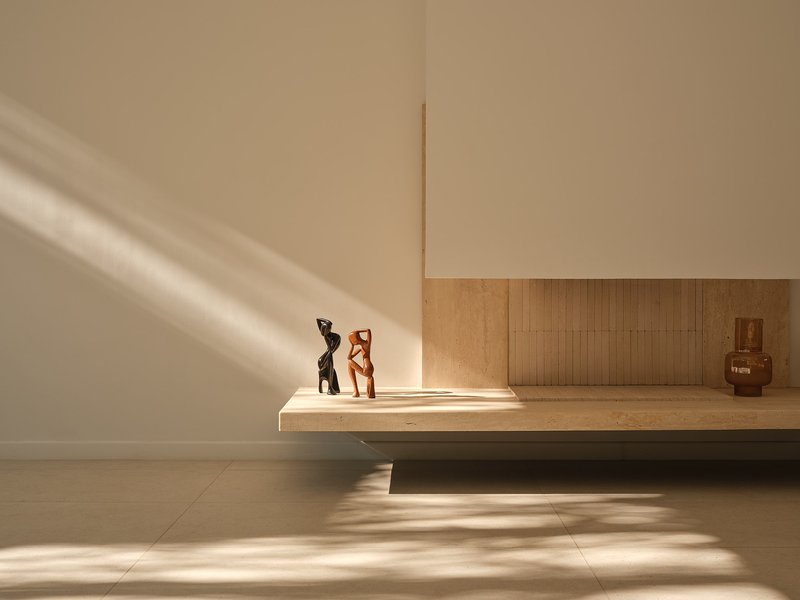 Low timber bench with sculptures lit by angular afternoon sunlight across pale floor tiles