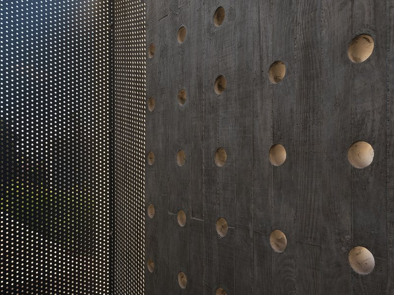Perforated metal screen beside board-formed concrete wall with exposed tie holes and green foliage beyond