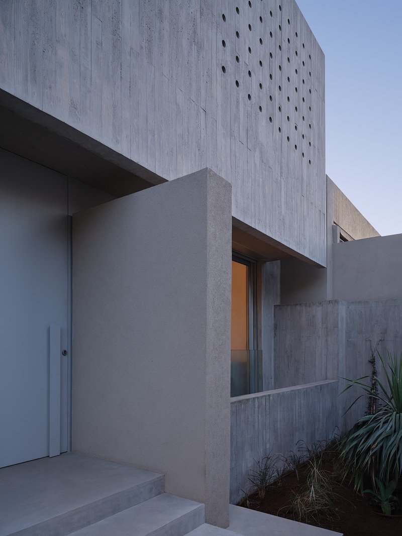 Concrete facade with scattered circular perforations at dusk beside planted beds with spiky grasses