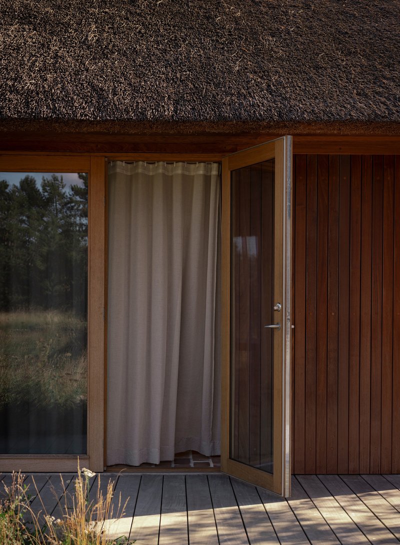 Timber deck threshold with open glass door and linen curtain under the thatched eave
