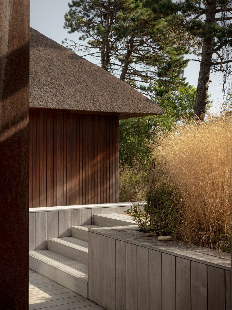 Weathered timber steps and bench beneath the thatched roof with pine trees and grasses