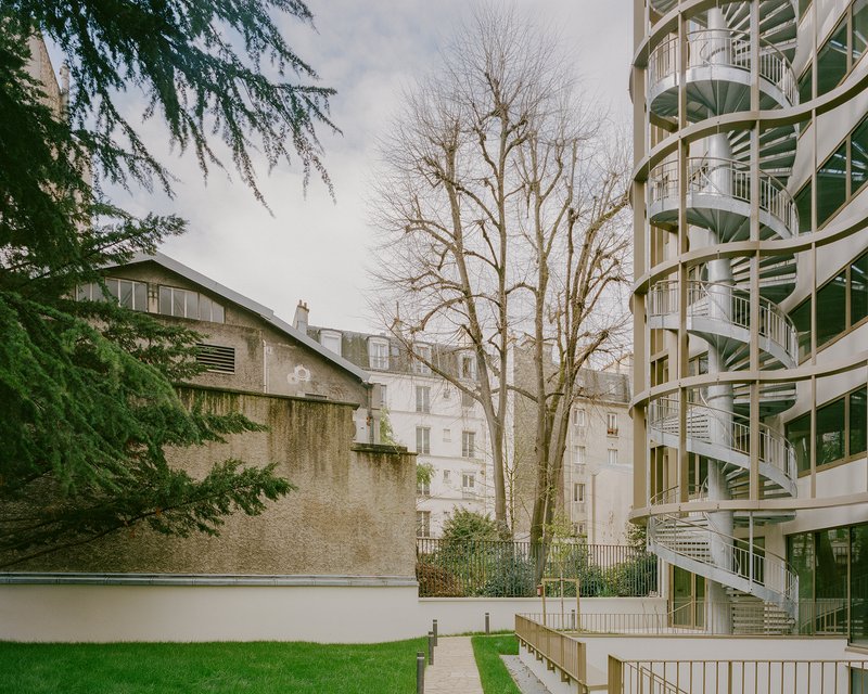 Curved facade with exposed spiral staircases beside a bare deciduous tree in a courtyard lawn