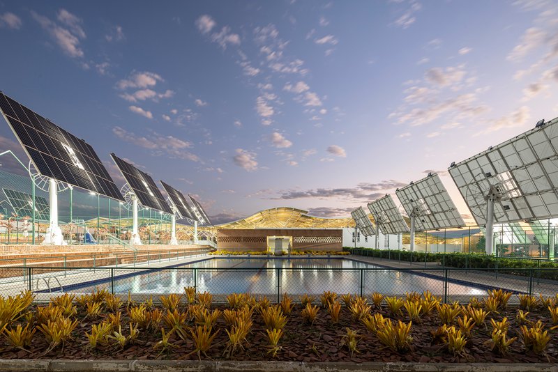 Reflecting pool with planted bed in foreground and solar panel canopies flanking a central pavilion at dusk