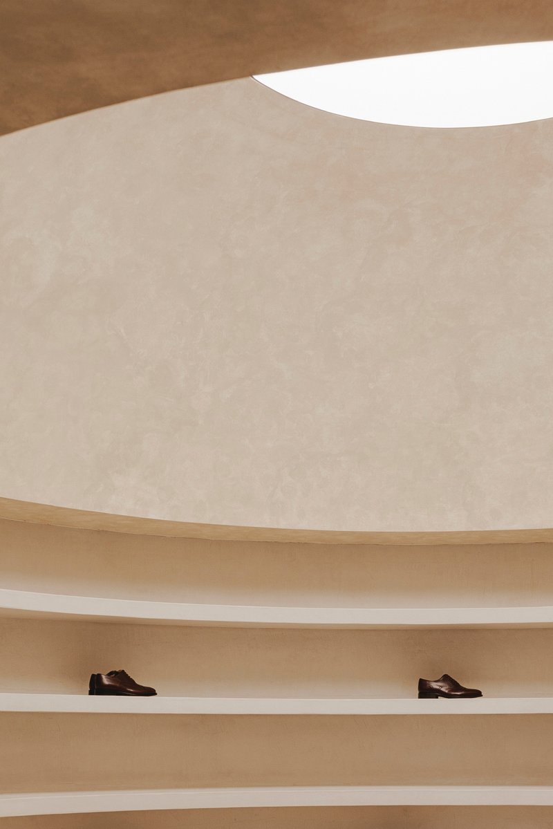 Curved plaster wall with circular skylight above continuous timber shelving holding two shoes