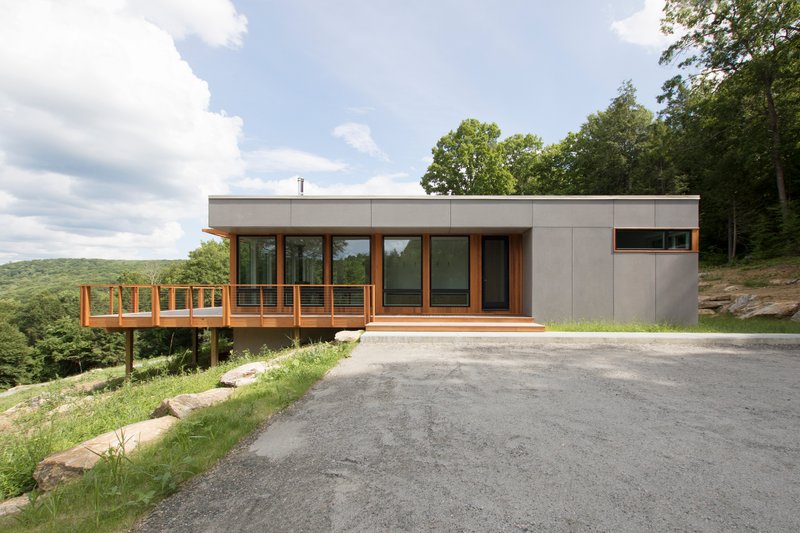 Street-facing view of the gray panel facade with cantilevered timber deck overlooking a forested hillside