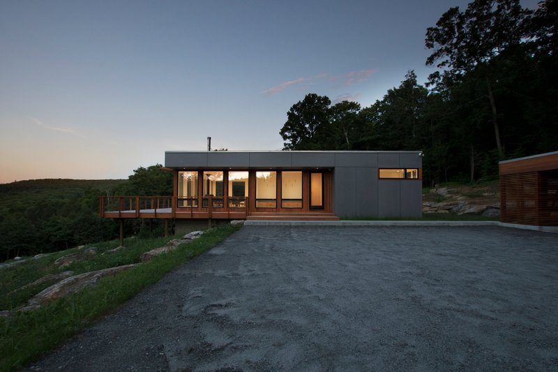 Grey-clad rectangular volume with full-height windows and timber deck overlooking forested hillside at twilight