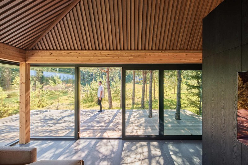 Interior view through floor-to-ceiling glazing to exterior terrace with timber slat ceiling above and figure standing outside