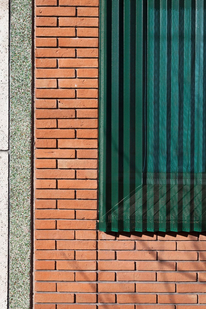 Detail of perforated green metal shutters meeting red brick wall with exposed aggregate concrete edge