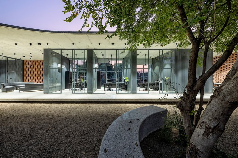 Glass facade beneath concrete overhang with existing mature tree shading a carved stone bench in gravel courtyard
