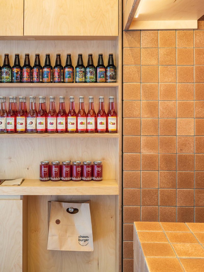 Open plywood shelving with bottled drinks beside orange tiled wall and takeaway bag below