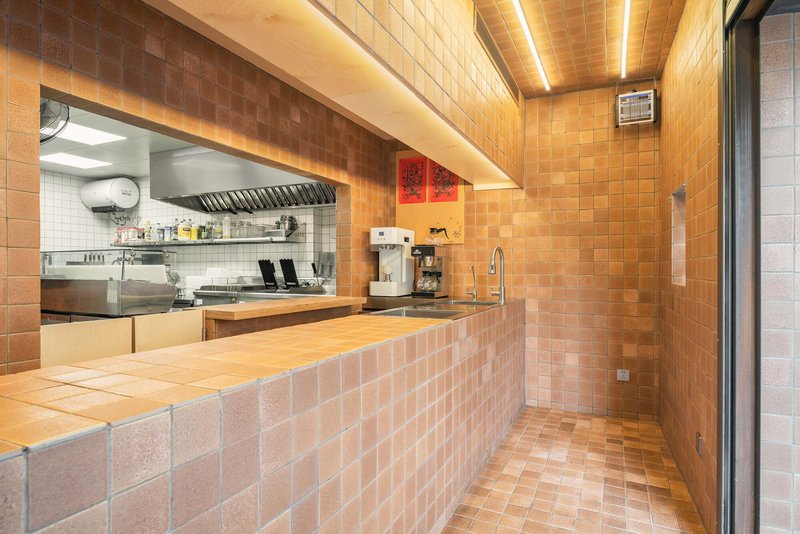 Interior service counter clad in terracotta tiles with linear ceiling lighting and kitchen view beyond