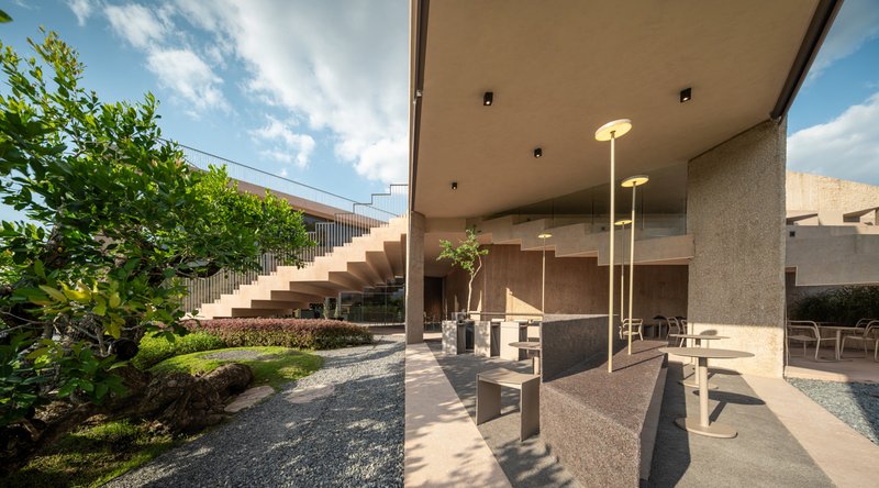 Covered terrace with concrete seating and exterior staircase ascending alongside planted gravel beds under blue sky