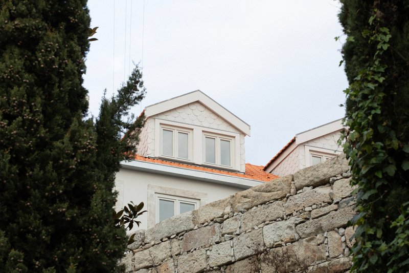 View of twin gabled volumes with white facades rising above a dry-stacked stone wall framed by tall cypress trees