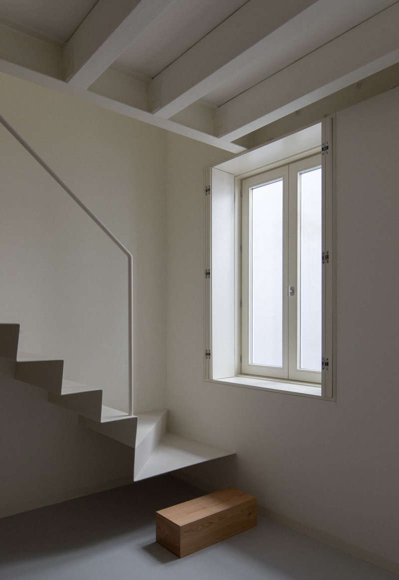 Interior staircase in white plaster with exposed ceiling beams and a casement window casting afternoon light