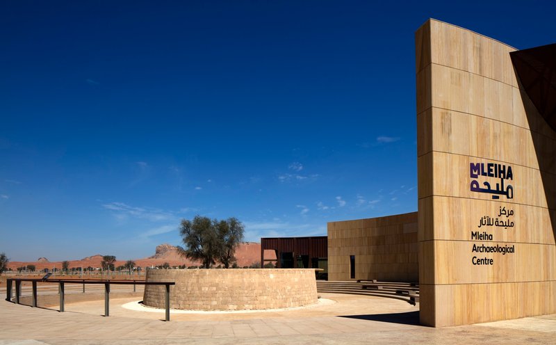 Wood-clad entrance tower with signage beside a curved stone wall and desert landscape beyond